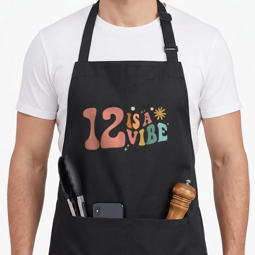 12th boho apron - Retro 12 Is A Vibe text in pink orange yellow green blue with flowers and stars — shown on a black cotton canvas apron against a white background
