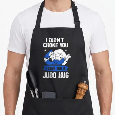 Fighter judo addict apron - I didn't choke you judo hug text with two grappling figures in white and blue — shown on a black cotton canvas apron against a white background