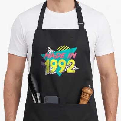 34th birthday apron - MADE IN 1992 text over teal triangle and white geometric shapes with black patterns — shown on a black cotton canvas apron against a white background