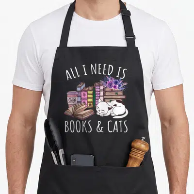 Book kitten cat apron - Sleeping white cat with stacked books and coffee cup under all I need is books and cats text — shown on a black cotton canvas apron against a white background