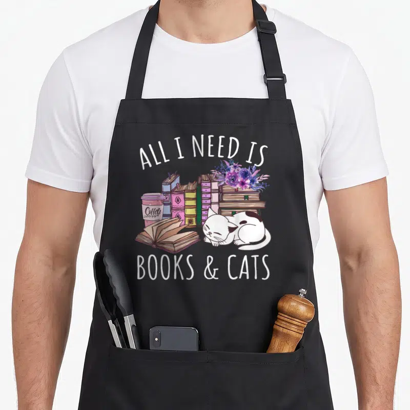 Book kitten cat apron - Sleeping white cat with stacked books and coffee cup under all I need is books and cats text — shown on a black cotton canvas apron against a white background
