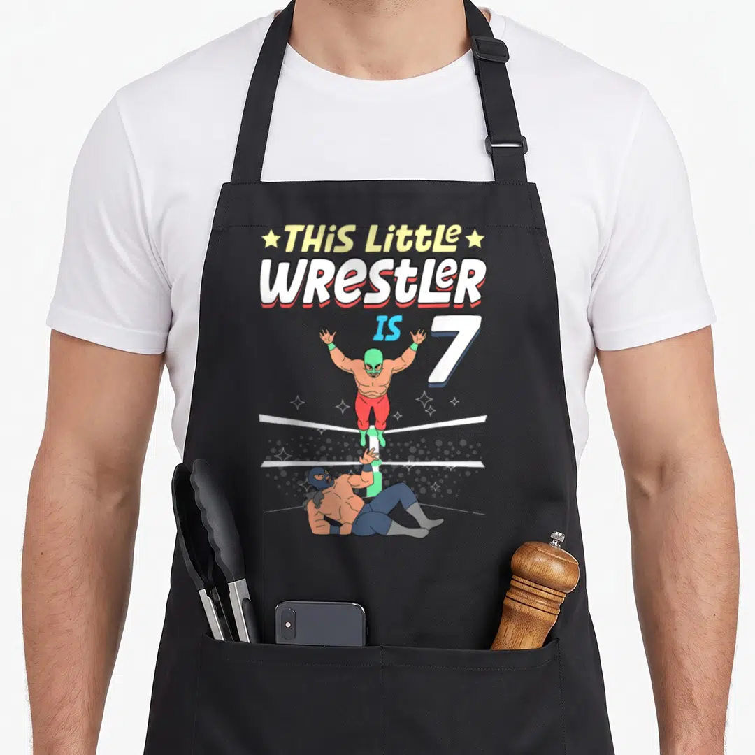 Wrestling birthday apron - This Little Wrestler is 7 text above green and blue masked wrestlers in a ring with white — shown on a black cotton canvas apron against a white background