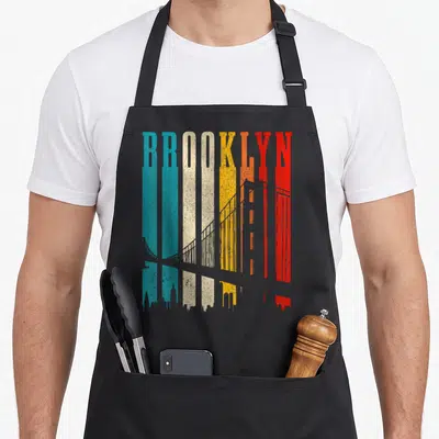 Bridge new york apron - Retro BROOKLYN text in colorful stripes with grey bridge and skyline silhouettes — shown on a black cotton canvas apron against a white background