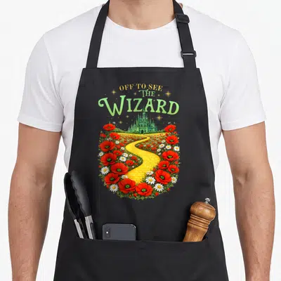 See road apron - OFF TO SEE THE WIZARD text above yellow brick road, emerald city, red poppies, white daisies — shown on a black cotton canvas apron against a white background