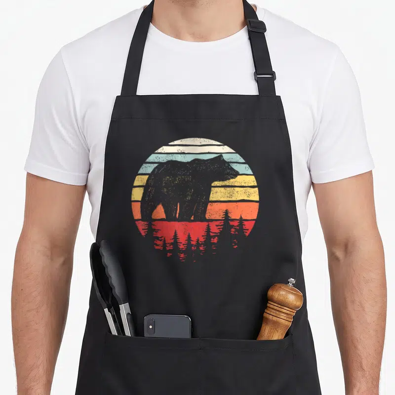 Catch bear apron - Grey bear silhouette against striped sunset with red water reflection and pine trees — shown on a black cotton canvas apron against a white background