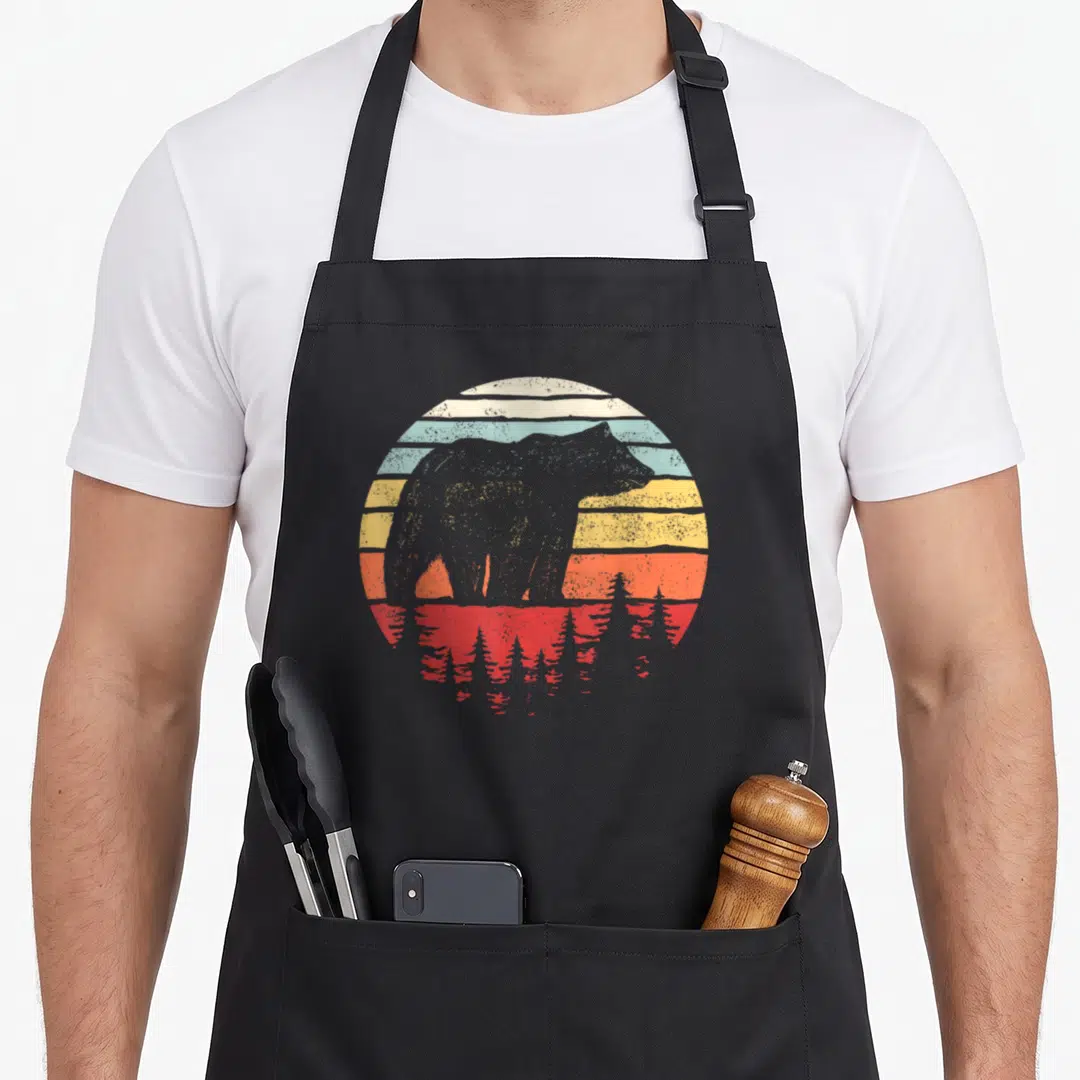 Catch bear apron - Grey bear silhouette against striped sunset with red water reflection and pine trees — shown on a black cotton canvas apron against a white background