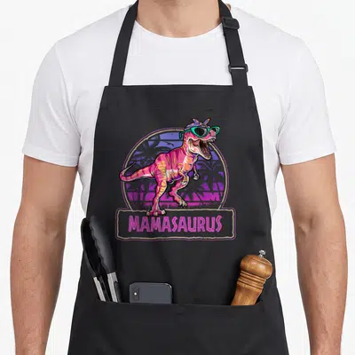 Saurus mama apron - Pink dinosaur with teal sunglasses and purple headband in front of palm trees with MAMASAURUS text — shown on a black cotton canvas apron against a white background