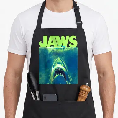 Surfacing novelty apron - Retro JAWS text in lime green above a shark rising from blue water under a swimmer — shown on a black cotton canvas apron against a white background