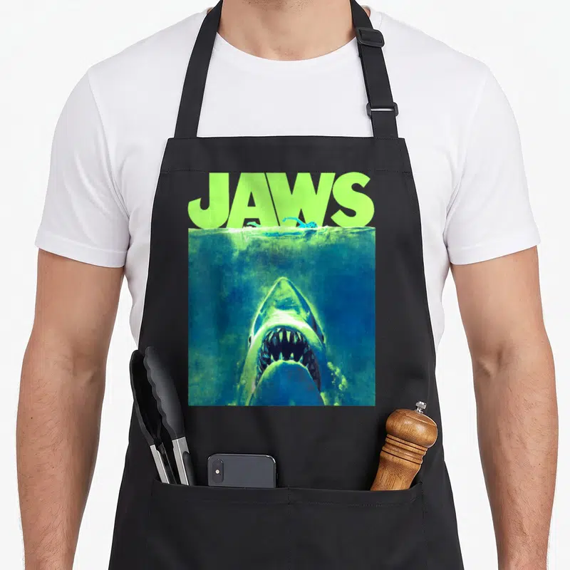 Surfacing novelty apron - Retro JAWS text in lime green above a shark rising from blue water under a swimmer — shown on a black cotton canvas apron against a white background