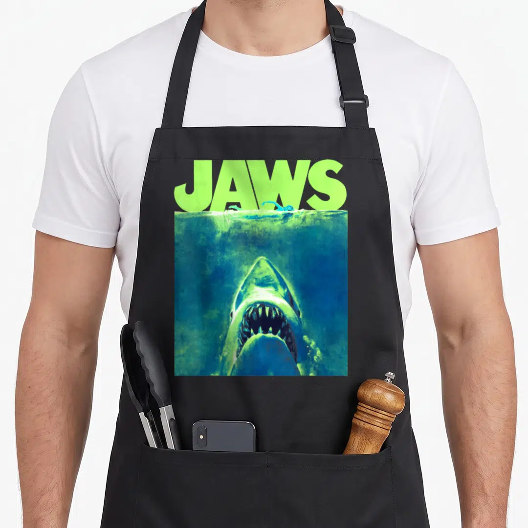 Surfacing novelty apron - Retro JAWS text in lime green above a shark rising from blue water under a swimmer — shown on a black cotton canvas apron against a white background