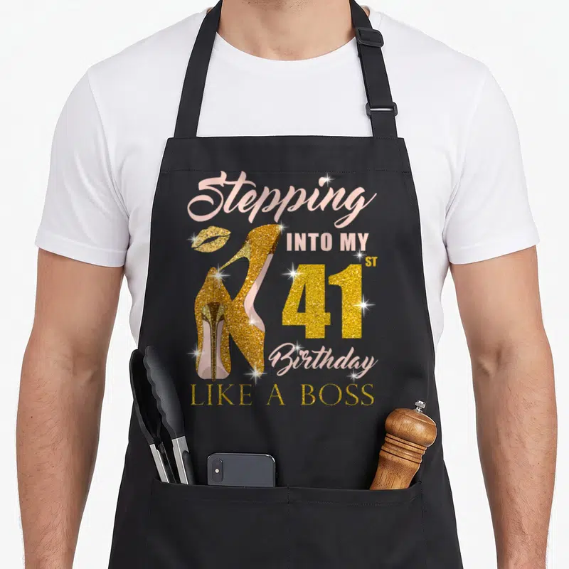 41st bbq boss apron - Gold glitter high heels and lips with Stepping Into My 41st Birthday Like A Boss text in pink — shown on a black cotton canvas apron against a white background