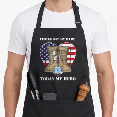Yesterday military apron - Tan combat boots and baby shoes over a US flag heart with dog tags — shown on a black cotton canvas apron against a white background