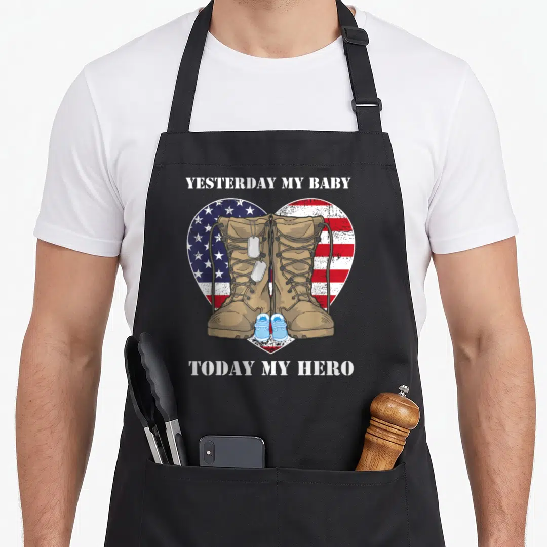 Yesterday military apron - Tan combat boots and baby shoes over a US flag heart with dog tags — shown on a black cotton canvas apron against a white background