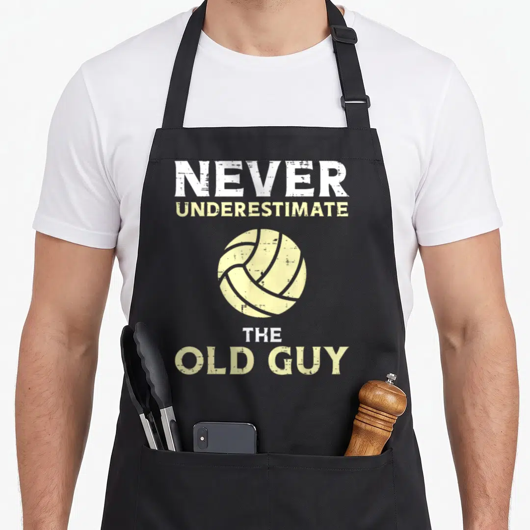 Volleyball grandpa apron - Stacked white and yellow text reading Never Underestimate The Old Guy above a distressed — shown on a black cotton canvas apron against a white background
