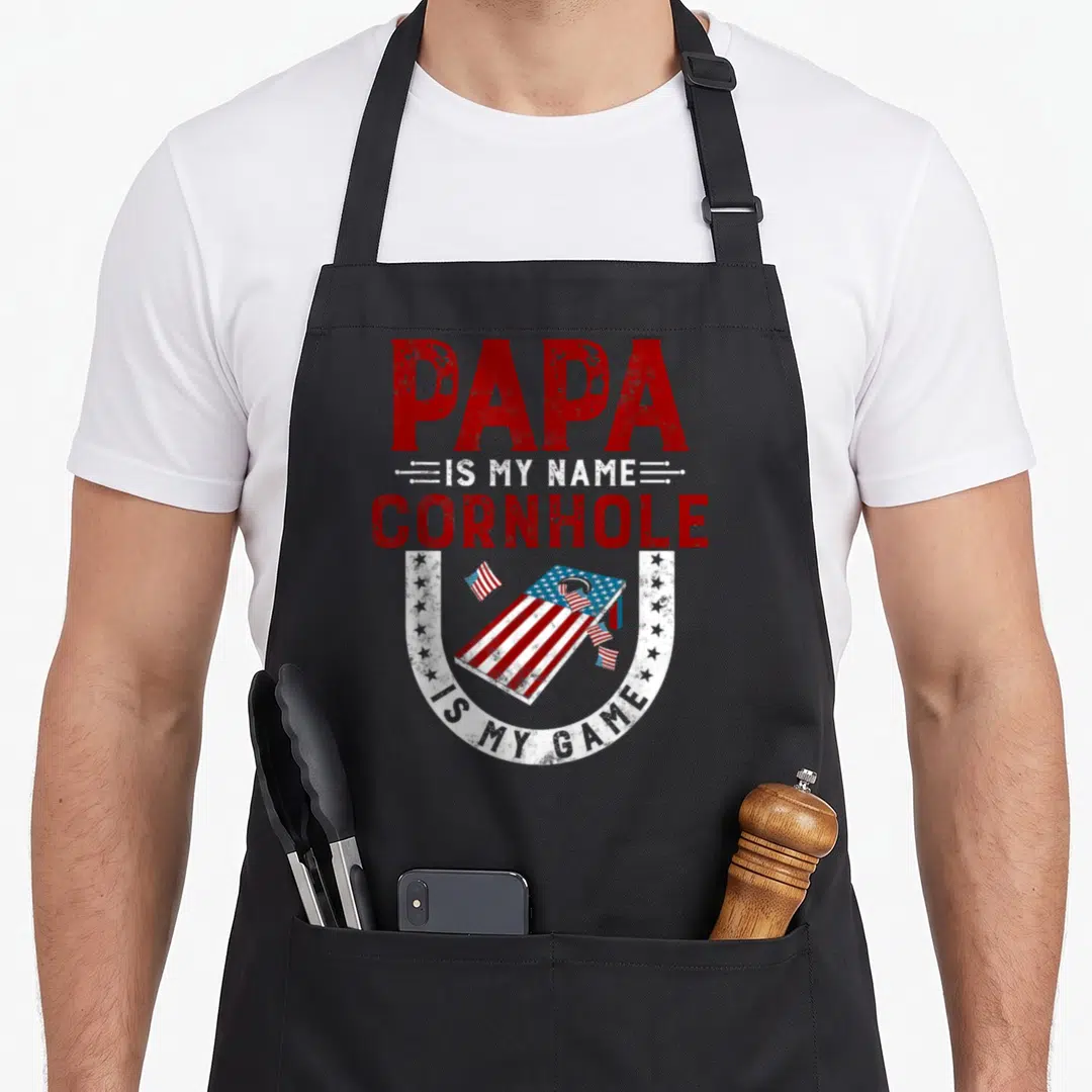 Cornhole papa apron - Distressed red white and blue text surrounding a US flag patterned cornhole board inside — shown on a black cotton canvas apron against a white background
