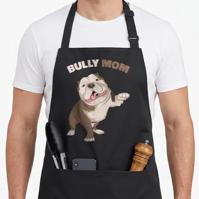 Bully dog mom apron - Cartoon bulldog with arched BULLY MOM text in brown, white, and pink — shown on a black cotton canvas apron against a white background
