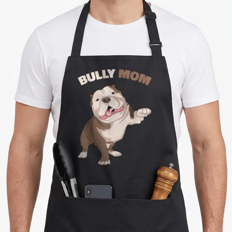 Bully dog mom apron - Cartoon bulldog with arched BULLY MOM text in brown, white, and pink — shown on a black cotton canvas apron against a white background