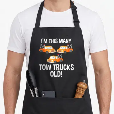Birthday tow truck apron - Three orange cartoon tow trucks in triangle with I'M THIS MANY TOW TRUCKS OLD text — shown on a black cotton canvas apron against a white background