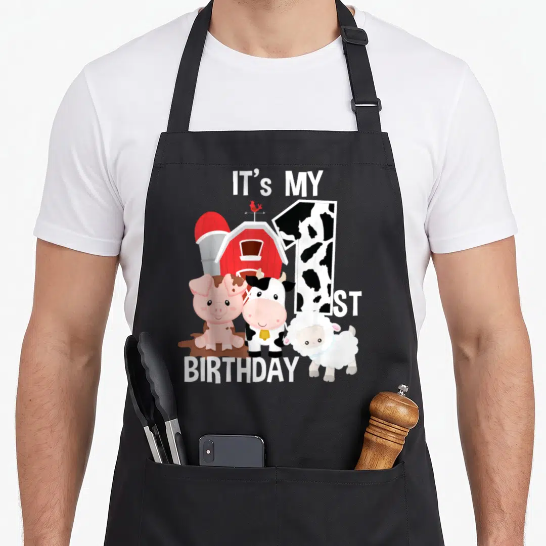 1st farm aprons - Cartoon pig cow sheep standing by red barn with cow print number 1 and white birthday text — shown on a black cotton canvas apron against a white background