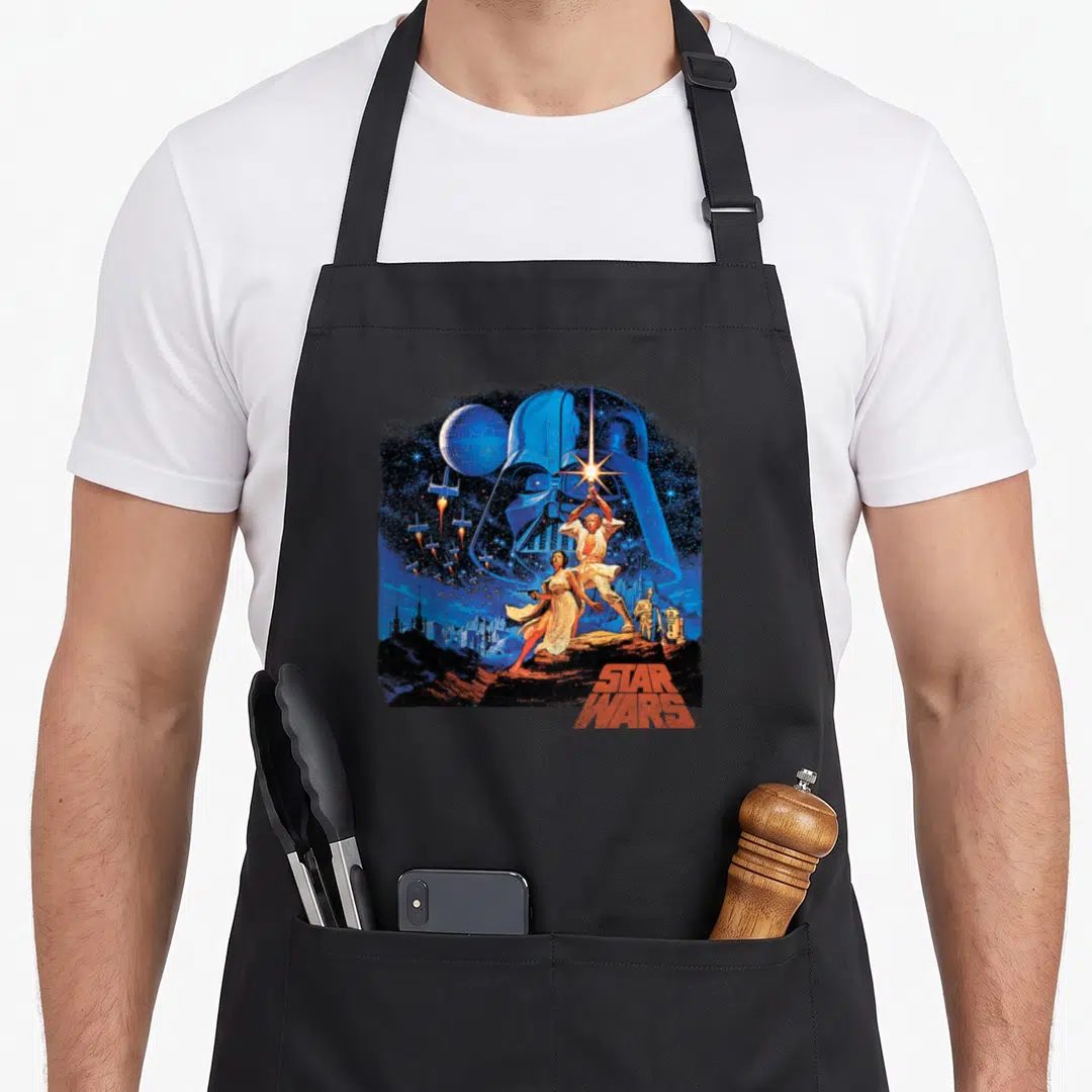 Poster new hope apron - Giant blue helmet, starships, mountain characters, red STAR WARS text — shown on a black cotton canvas apron against a white background