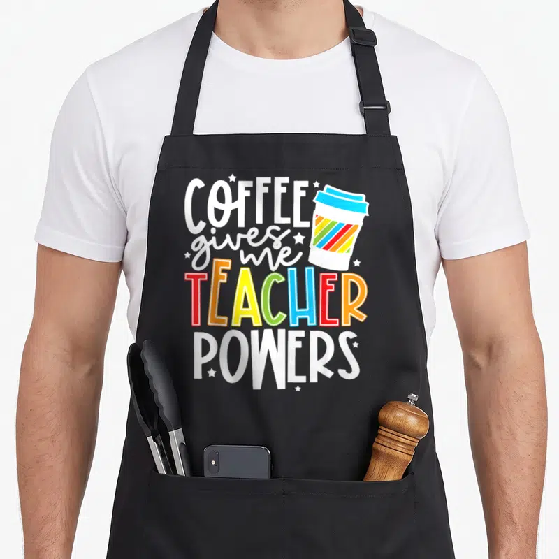 Coffee teacher apron - COFFEE gives me TEACHER POWERS text with rainbow striped cup and white stars — shown on a black cotton canvas apron against a white background