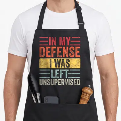 Unsupervised remaining apron - Stacked retro text In My Defense I Was Left Unsupervised in red orange yellow blue white — shown on a black cotton canvas apron against a white background