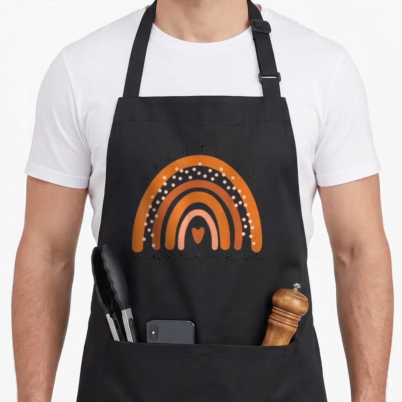 Infertility april apron - Rainbow in orange peach and brown with INFERTILITY text and heart — shown on a black cotton canvas apron against a white background