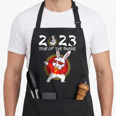 Funny chinese rabbit apron - Cartoon white rabbit in red jacket and sunglasses dabbing under 2023 text and YEAR — shown on a black cotton canvas apron against a white background