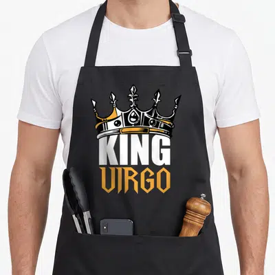 Virgo king apron - Cartoon crown sitting atop stacked KING and VIRGO text in black orange and white — shown on a black cotton canvas apron against a white background