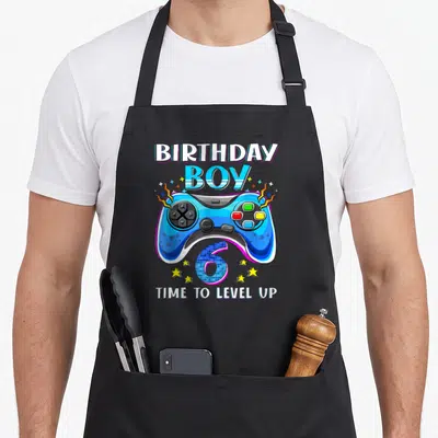 Vintage birthday video game apron - Blue game controller with BIRTHDAY BOY 6 TIME TO LEVEL UP text in cartoon style — shown on a black cotton canvas apron against a white background