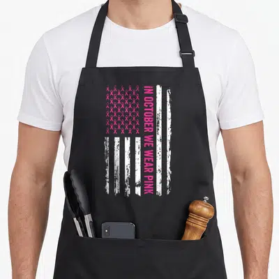 October bow apron - Distressed USA flag pattern with pink ribbon stars and vertical text reading IN OCTOBER WE WEAR PINK — shown on a black cotton canvas apron against a white background