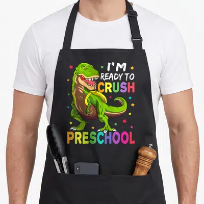 Ready preschool apron - Green T-Rex with yellow backpack and rainbow text I'm ready to crush preschool — shown on a black cotton canvas apron against a white background