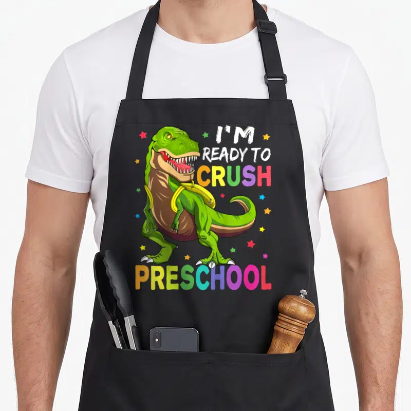 Ready preschool apron - Green T-Rex with yellow backpack and rainbow text I'm ready to crush preschool — shown on a black cotton canvas apron against a white background