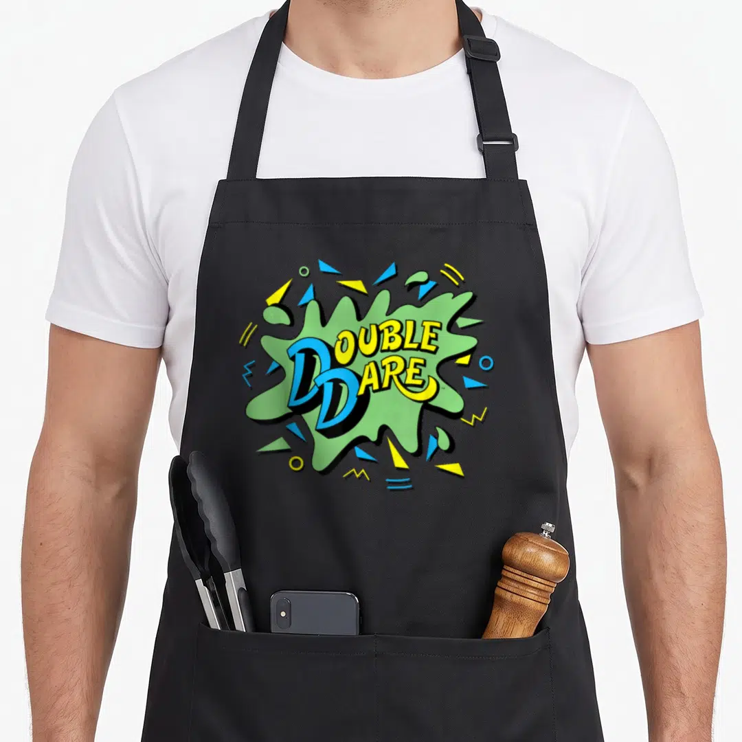 Dare double apron - DOUBLE DARE text in yellow and blue on a green splat with geometric shapes — shown on a black cotton canvas apron against a white background