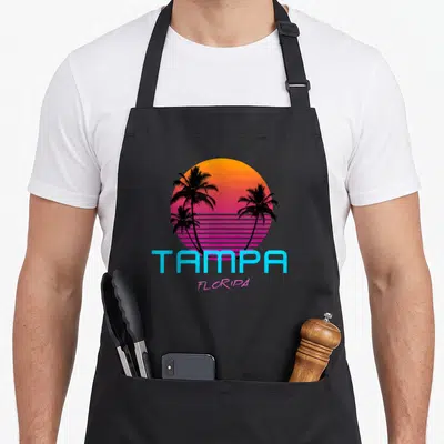 Tampa 80s apron - Gradient sun with three black palm trees and blue TAMPA pink FLORIPA text — shown on a black cotton canvas apron against a white background