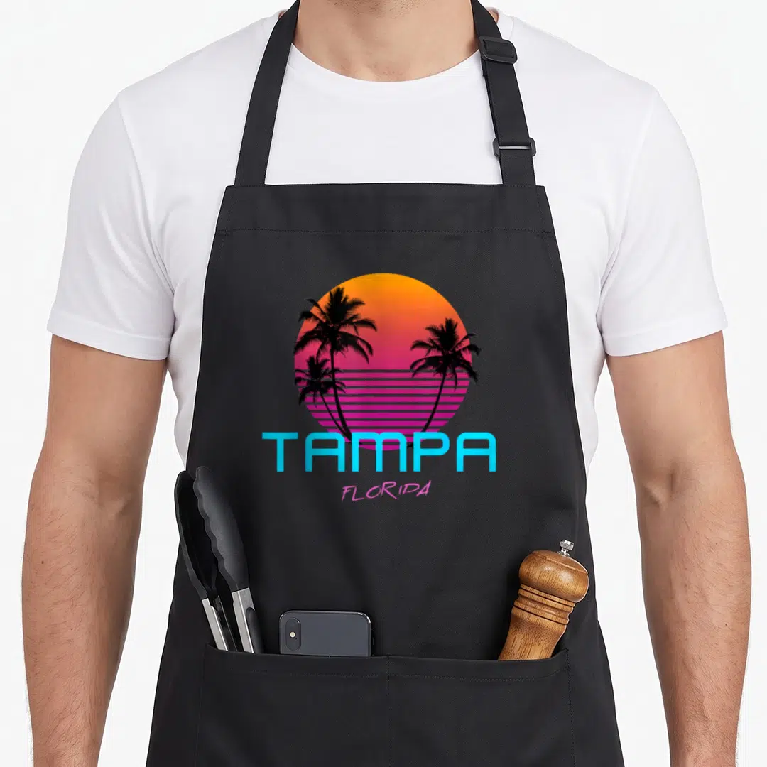 Tampa 80s apron - Gradient sun with three black palm trees and blue TAMPA pink FLORIPA text — shown on a black cotton canvas apron against a white background