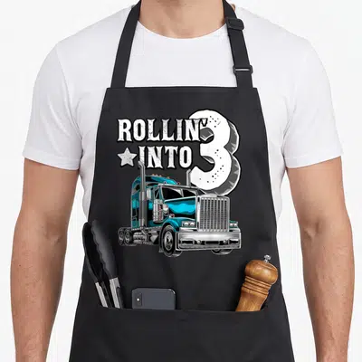 3rd semi truck apron - Blue cartoon big rig truck with ROLLIN INTO 3 text and star graphic — shown on a black cotton canvas apron against a white background