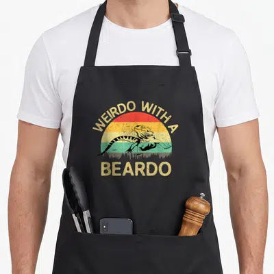 Weirdo beardo dragon apron - Retro bearded dragon standing before striped sun with WEIRDO WITH A BEARDO text — shown on a black cotton canvas apron against a white background