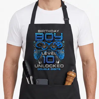 Unlocked 10th birthday gaming aprons - Blue game controller with BIRTHDAY BOY LEVEL 10 UNLOCKED DOUBLE DIGITS pixelated — shown on a black cotton canvas apron against a white background