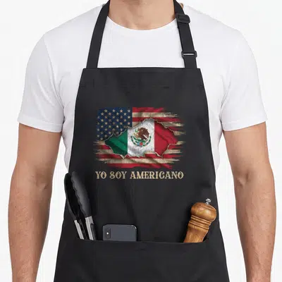 Americano spanish aprons - Distressed US flag tearing open to show Mexican flag with text YO SOY AMERICANO below — shown on a black cotton canvas apron against a white background