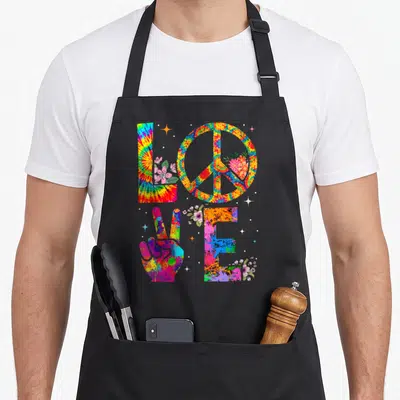 60s tie dye apron - Tie-dye LOVE text with peace circle, hand gesture, and flowers in red yellow blue green pink — shown on a black cotton canvas apron against a white background