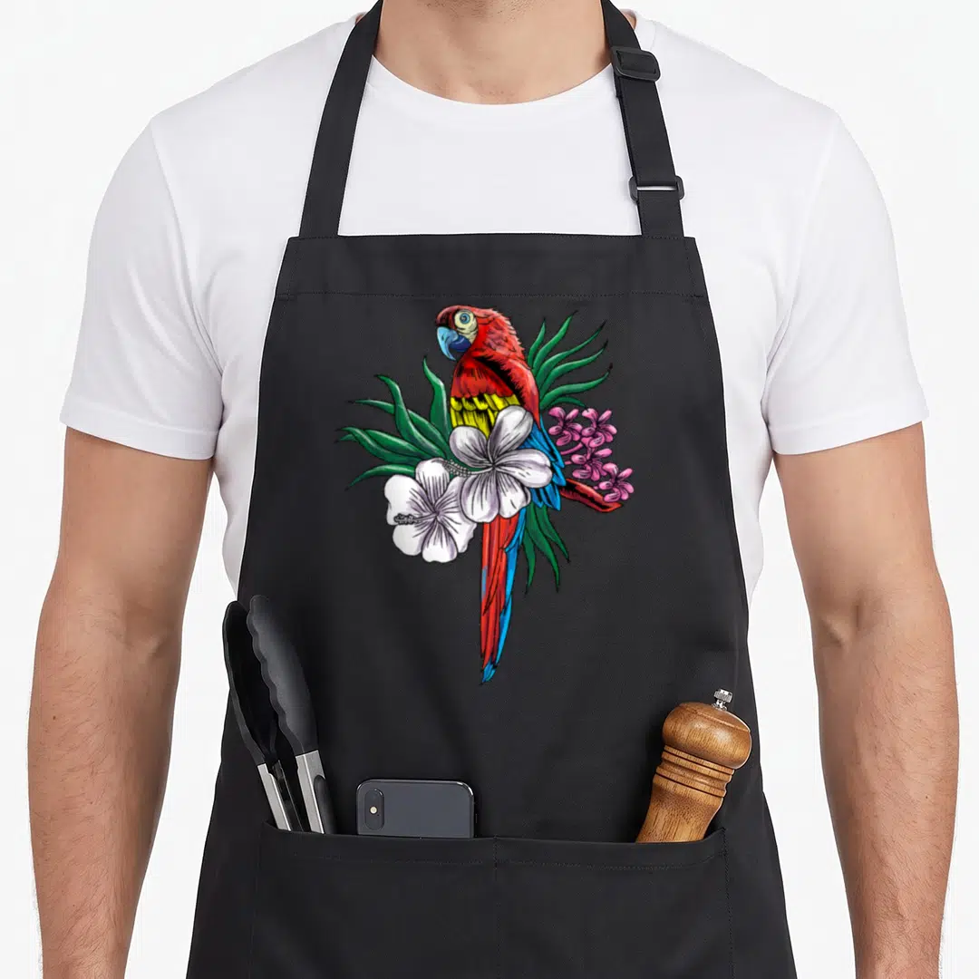 Tropical bird apron - Cartoon macaw with red blue yellow feathers surrounded by green leaves white hibiscus and pink — shown on a black cotton canvas apron against a white background