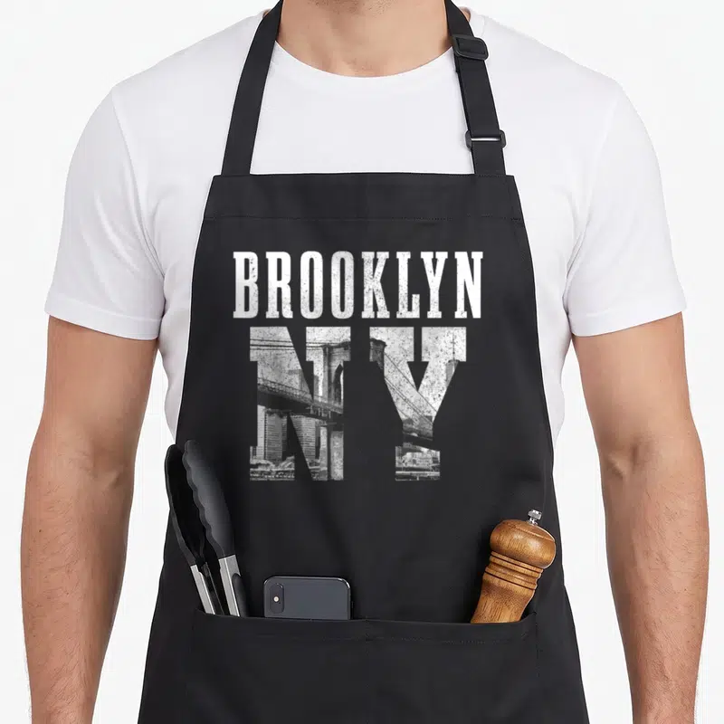 Retro brooklyn new york apron - Distressed white BROOKLYN text above grey NY letters containing a bridge and skyline — shown on a black cotton canvas apron against a white background