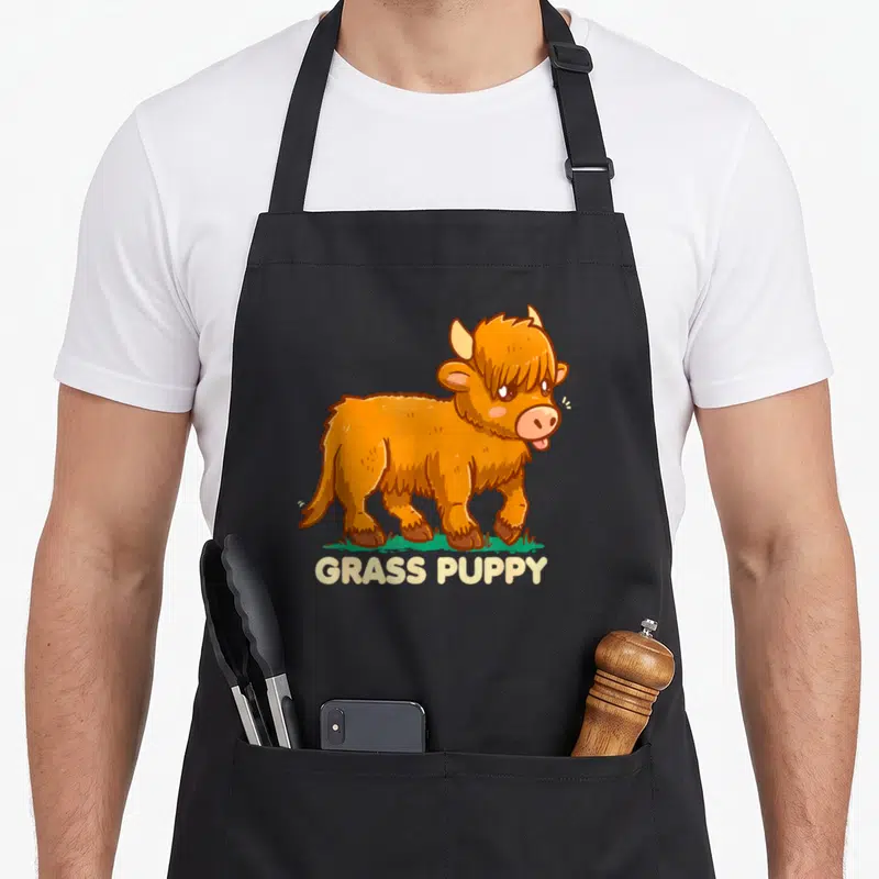 Grass highland cow apron - Orange cartoon highland cow standing on green grass with GRASS PUPPY text below — shown on a black cotton canvas apron against a white background