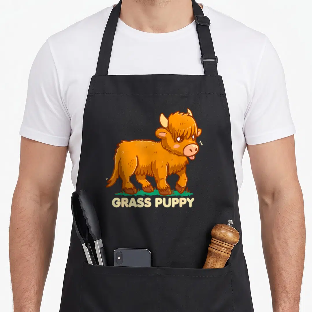 Grass highland cow apron - Orange cartoon highland cow standing on green grass with GRASS PUPPY text below — shown on a black cotton canvas apron against a white background