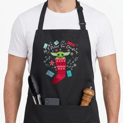 Funny general christmas apron - Grogu sitting in red stocking surrounded by blue gifts snowflakes and white arched text — shown on a black cotton canvas apron against a white background