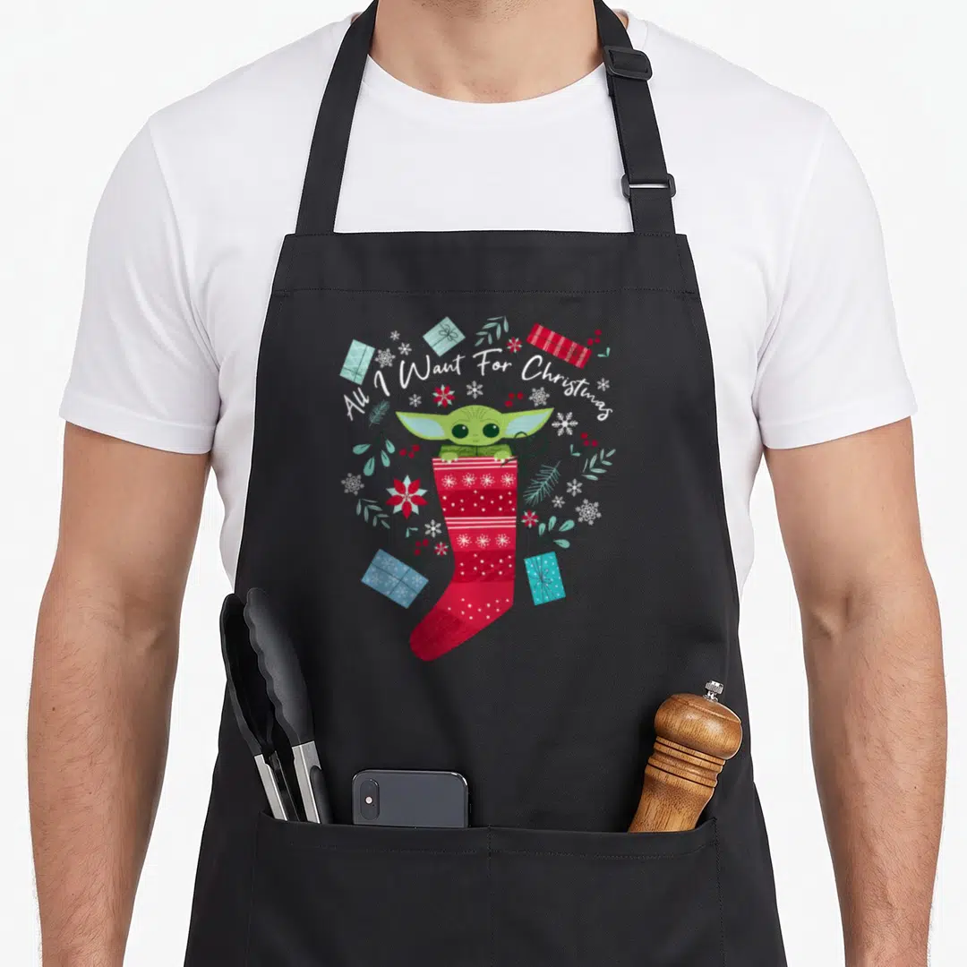 Funny general christmas apron - Grogu sitting in red stocking surrounded by blue gifts snowflakes and white arched text — shown on a black cotton canvas apron against a white background