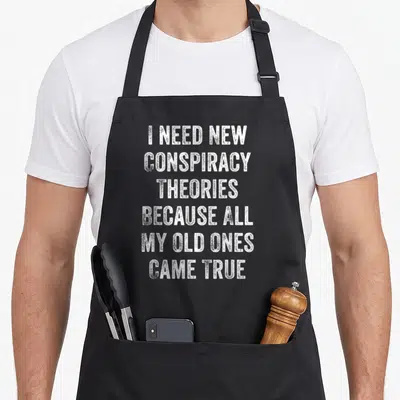 Conspiracy theorist apron - Distressed white text I NEED NEW CONSPIRACY THEORIES BECAUSE ALL MY OLD ONES CAME TRUE — shown on a black cotton canvas apron against a white background