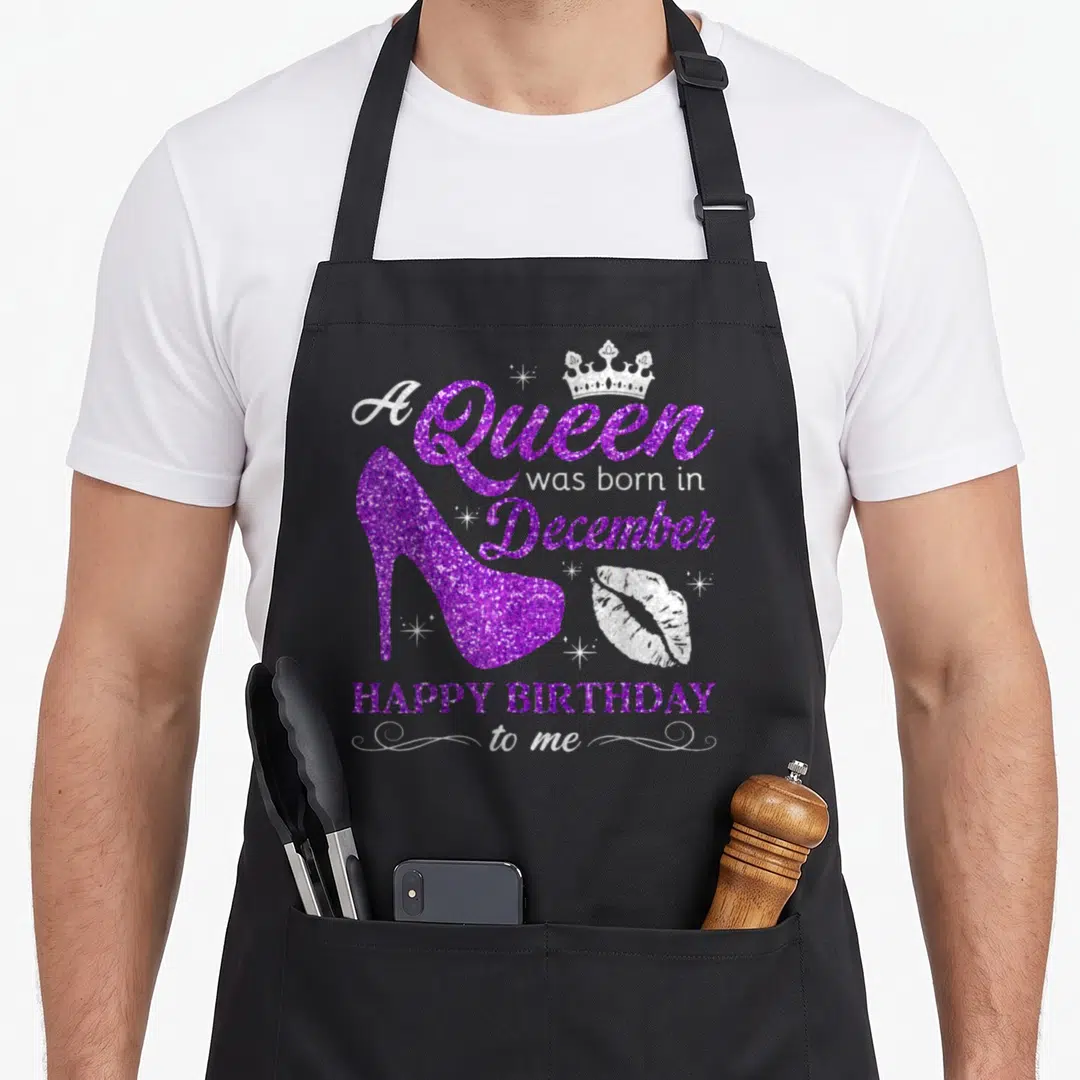 Funny queen december birthday apron - Purple glitter high heel shoe with crown and A Queen was born in December text — shown on a black cotton canvas apron against a white background