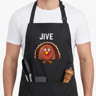 Funny dinner thanksgiving apron - Cartoon turkey in brown red yellow white below white text JIVE — shown on a black cotton canvas apron against a white background